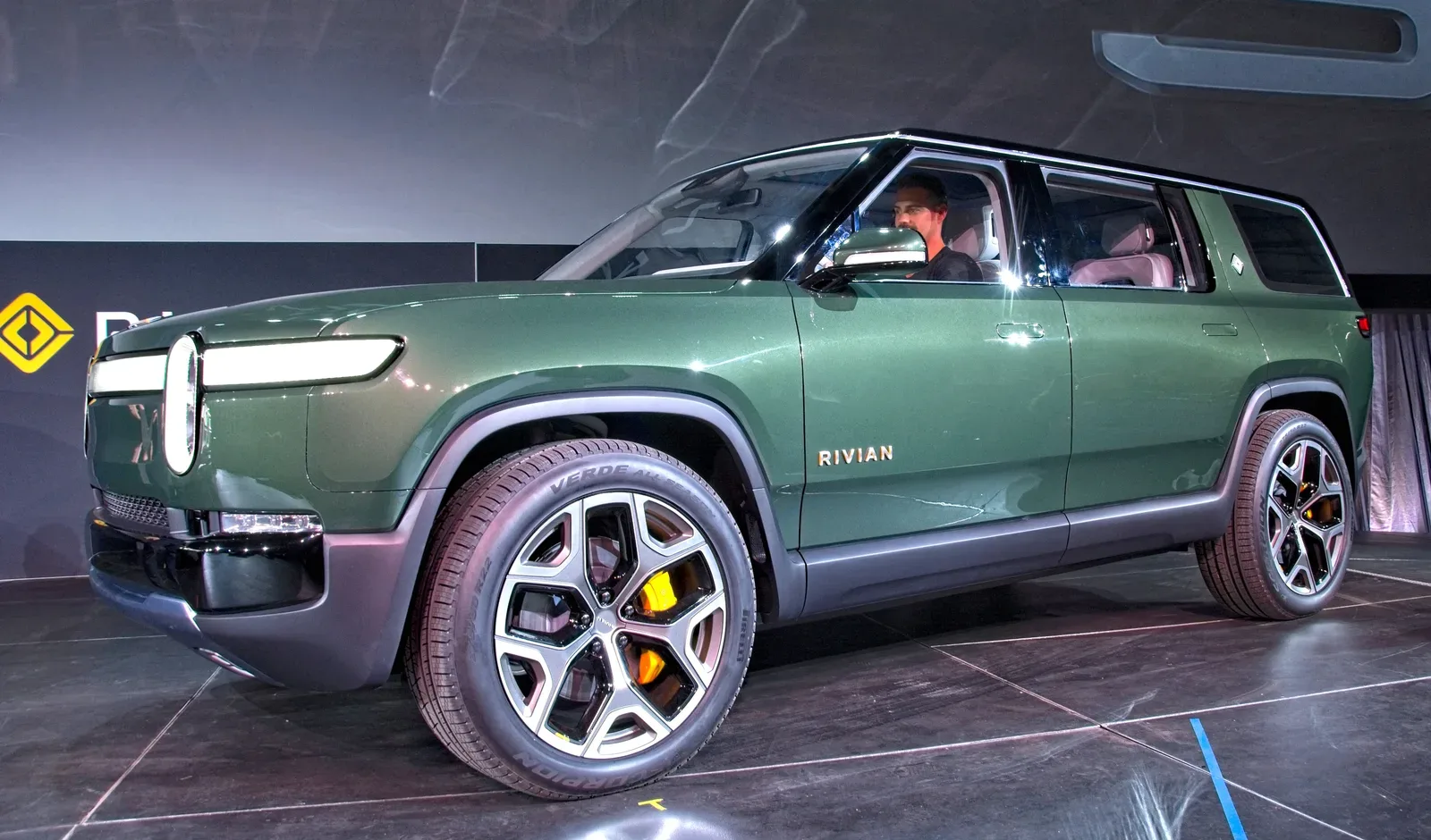 Un SUV eléctrico Rivian verde expuesto en un evento de presentación con conductor al volante