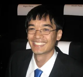 Retrato de un hombre sonriente con gafas, traje oscuro y corbata azul, en un acto.