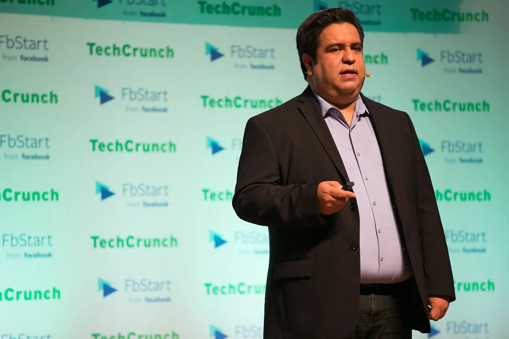 Hombre con micrófono de diadema habla en escenario ante fondo con logos de TechCrunch y FbStart
