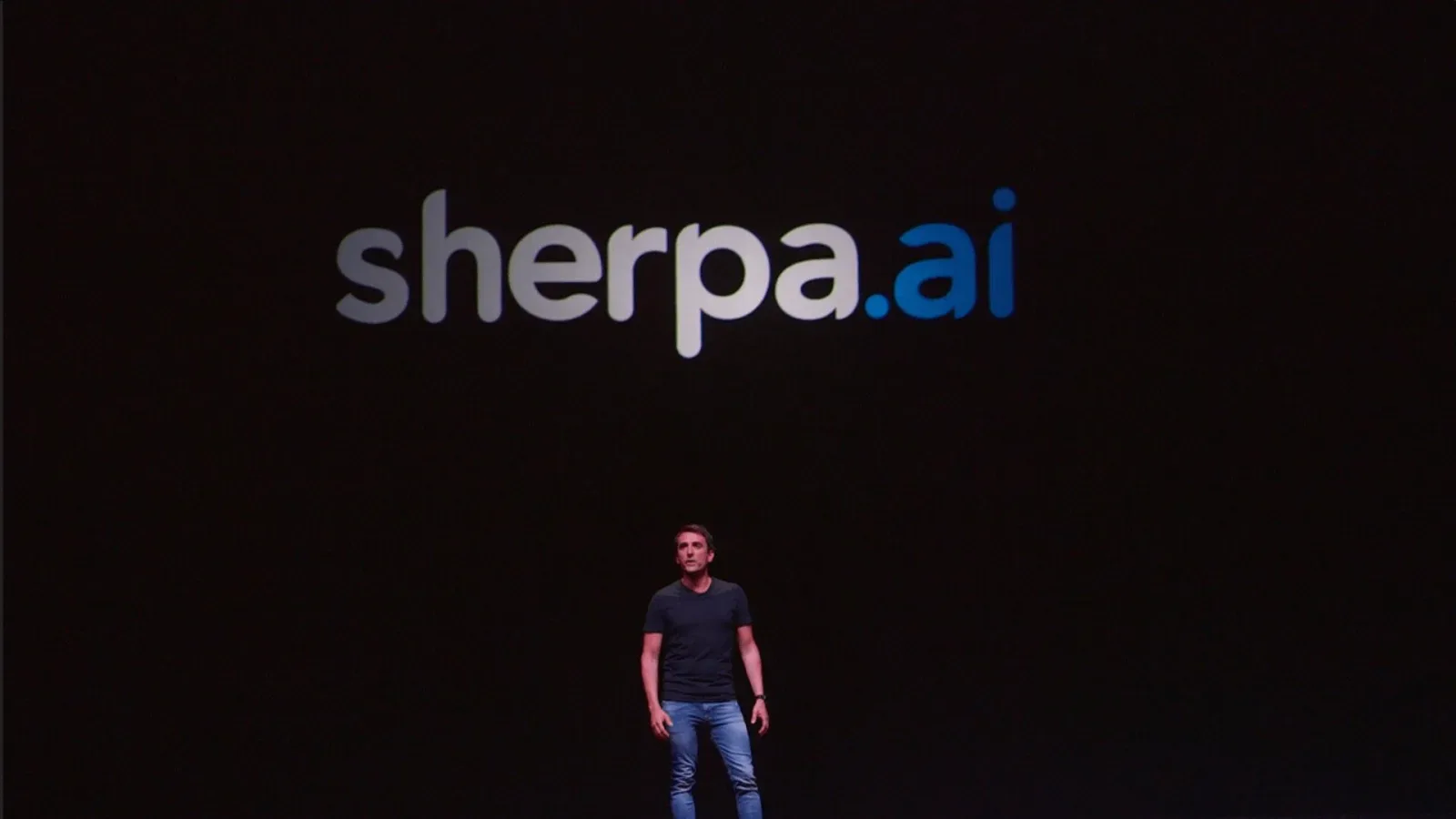 Un hombre presenta en un escenario bajo el logotipo de Sherpa.ai