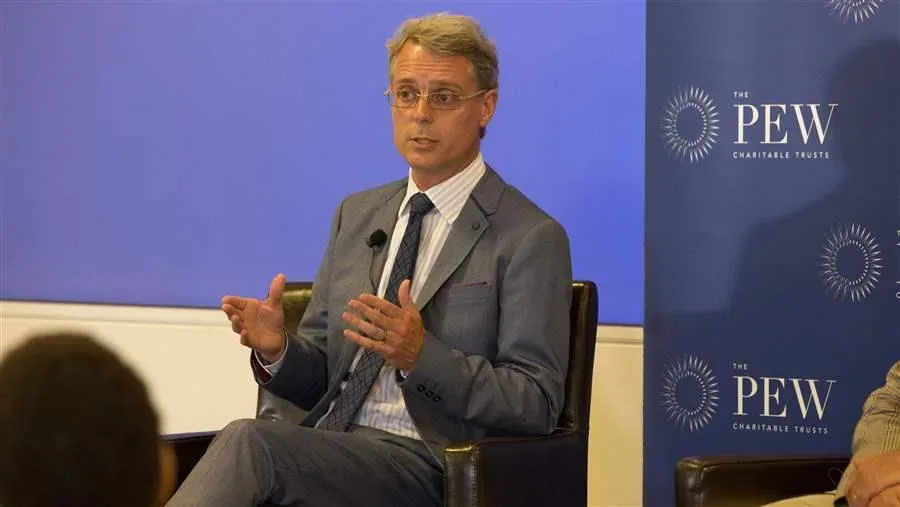 Hombre con traje gris habla sentado ante panel de Pew Charitable Trusts