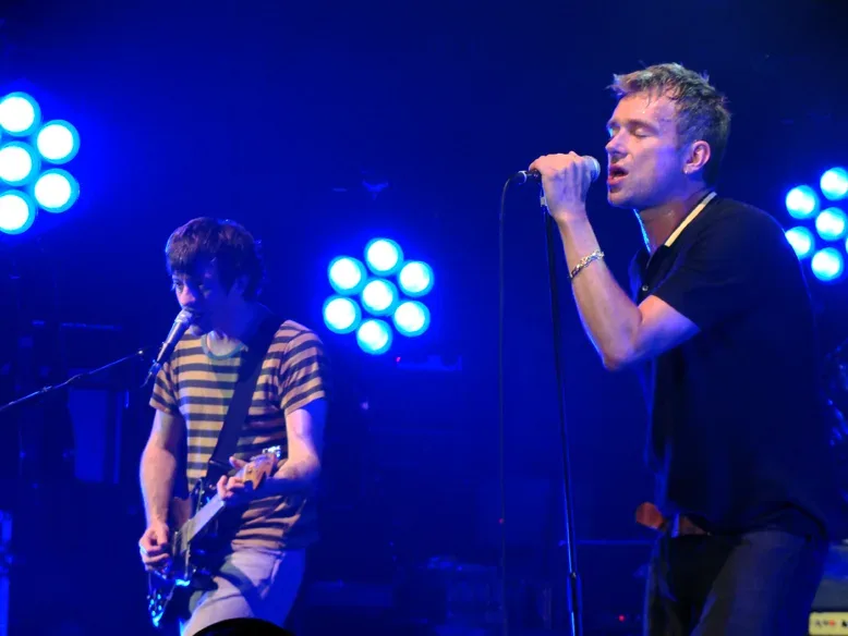 Graham Coxon toca la guitarra junto a un cantante durante un concierto en directo.