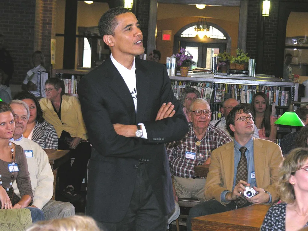 Barack Obama de pie, brazos cruzados, habla ante una audiencia en una biblioteca
